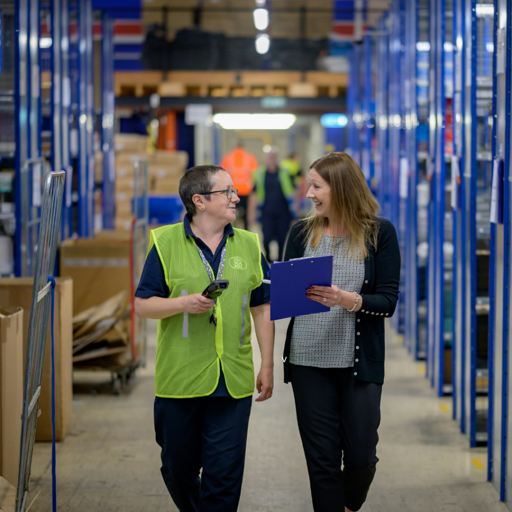 Two women walking through a warehouse and talking. One is holding a clipboard.