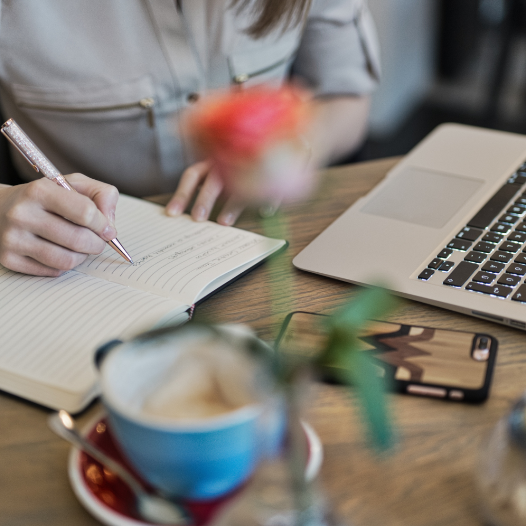 Person Writing On A Notebook Beside Macbook