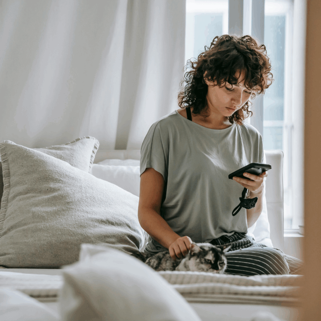 Serious young ethnic lady reading message and petting cat on bed